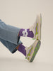 A person wearing purple and green socks, showcasing the UNDO x MUSTIQUE SNEAKERS.