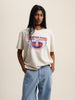 The model dons a white t-shirt featuring a skateboard image. The t-shirt is the Mustique Hotel T-shirt in ecru.